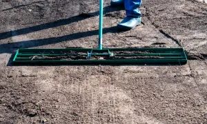 Osoba w niebieskich spodniach i gumowcach wyrównuje ziemię pod trawnik za pomocą zielonej równiarki.