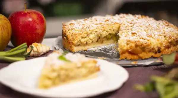 Krucha szarlotka z kawałkami kwaśnych jabłek, posypana cukrem pudrem. Obok leżą świeże jabłka i mały rogalik.