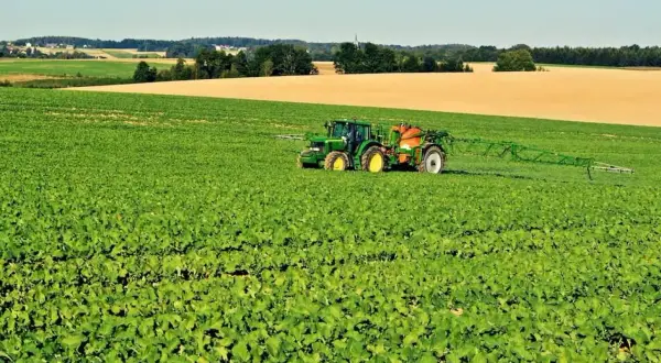 Zielony traktor z opryskiwaczem na polu rzepaku. W tle pola i wieś. Jakby uderzenie **inazuma** w krajobraz.