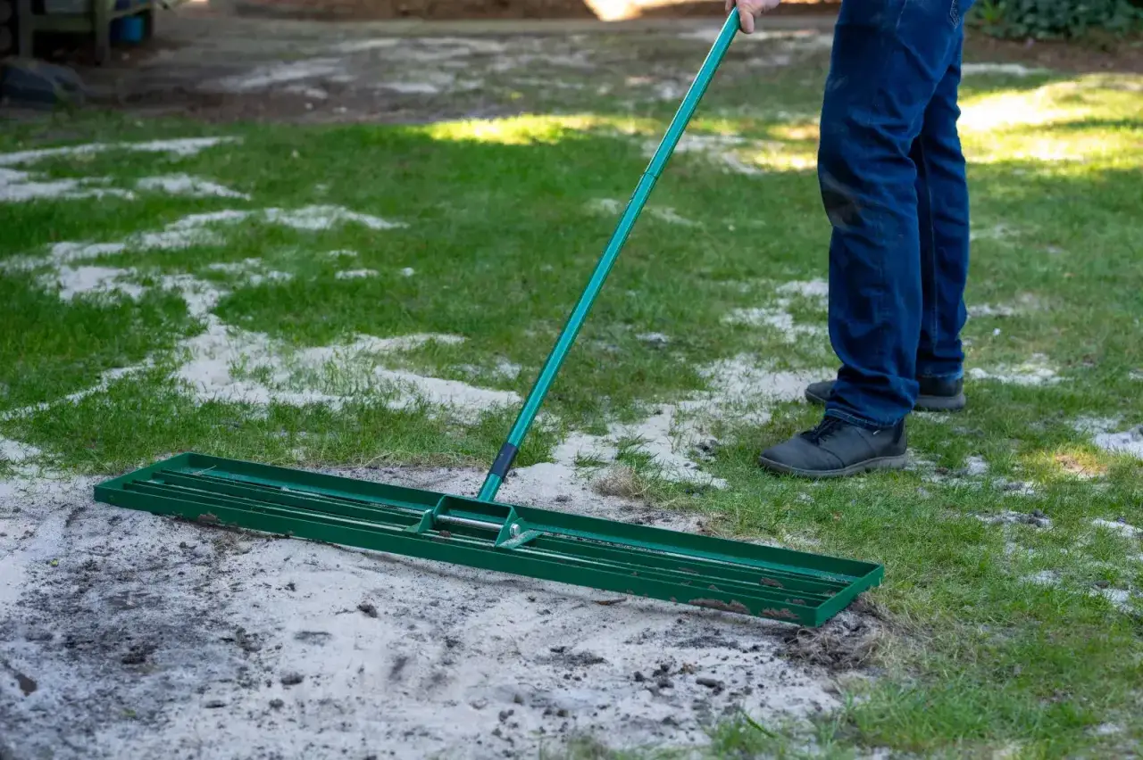 Mężczyzna wykonuje piaskowanie trawnika, rozprowadzając piasek za pomocą zielonego narzędzia.